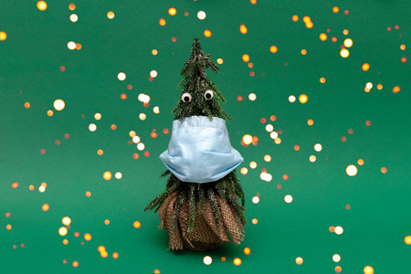 Christmas tree with blue facial medical protective mask and googly eyes on green background, bokeh style, celebration with lockdown due to covidの写真素材