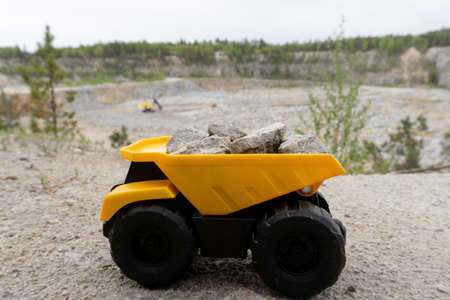 Closeup at small yellow car toy, mining truck with stones at the hill at the background of quarry. Stones and trees at background and blue sky.の写真素材