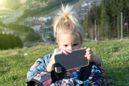 Cute little young blond caucasian toddler laying at green grass and looking at mobile phone with interested face, summer time, modern digital generation involved in mobile games, sun shines, outdoorの写真素材