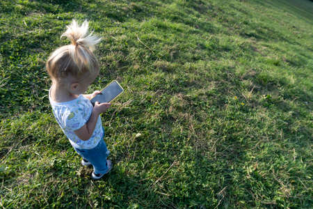 Cute caucasian blond little girl playing or learning at mobile phone with black screen, staying at green grass at summer, leisure time at nature of modern children, view from the top.の写真素材