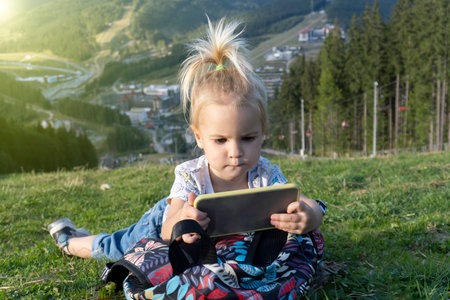 Cute little caucasian kid laying at green grass and looking at mobile phone with interested face, summer time, modern digital generation involved in mobile games.の写真素材