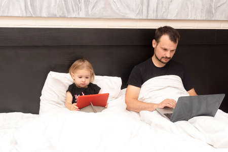 Caucasian young father is working from home with laptop in bed and caucasian little cute kid is near, watching cartoons, white linen, isolated period at lockdown with covid, remote work concept.の写真素材
