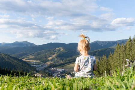 Cute little young child sitting at green grass at the top of the hill with the view of mountains at summer, blue sky with clouds, view from the back.の写真素材