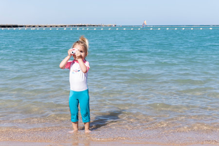 Small cute caucasian child is taking photo at children little camera near blue sea at summer vacation with family, happy memory shots, small beginner in photography.の写真素材