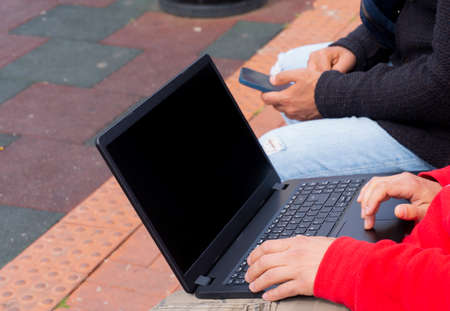 Two man sit on bench outdoor and using modern technology, laptop and mobile phone, close up, only hands and devices.の写真素材