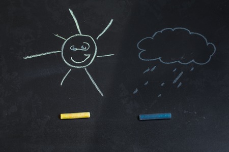 colored chalks on a black blackboard with drawings of a sun and a cloudの写真素材