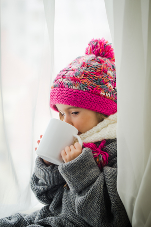 Cute little girl in fashionable winter clothes with a knitted hat and warm sweater. Holding in his hand a white Cup of tea.の写真素材