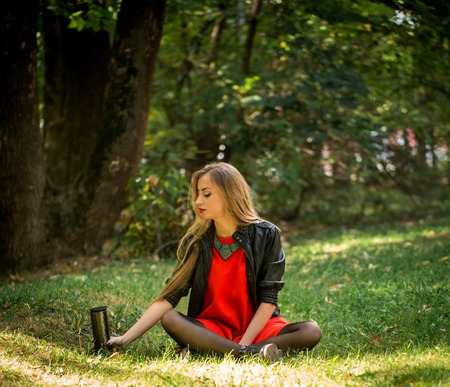 Beautiful girl with long hair in red dress and leather jacket sitting on the grass and holding a thermosの写真素材