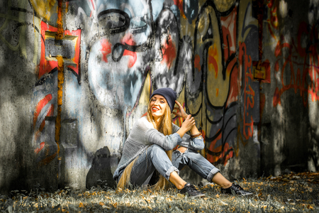 Fashionable girl in a hat with a long blond hair in stylish clothes standing on a background of graffitiの写真素材