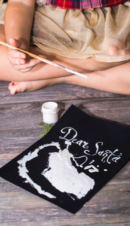 Beautiful cute little girl writes letter to Santa near Christmas decoration on the wooden floorの写真素材