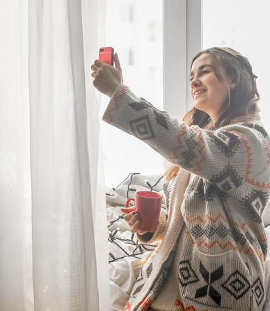 A cozy Cup of tea at a cute girl with a phone in a warm knitted sweater against the windowの写真素材