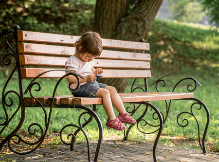 Little cute girl in the park on the bench with the phoneの写真素材