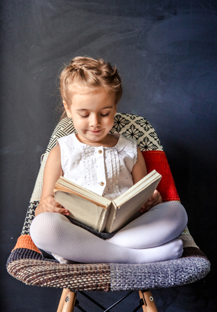 little girl sitting on a chair and reading a bookの写真素材