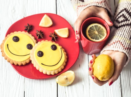 red Cup of hot tea with lemon in hands of the girl in the sweater,on a white wooden background red and a plate of cookies,the concept of hot drinksの写真素材