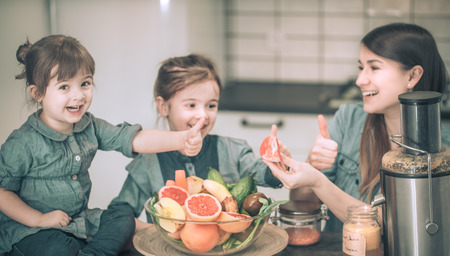mother with two lovely daughters prepares fresh fruit juice in the kitchen, the concept of a healthy baby food and a happy familyの写真素材