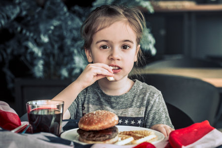 little girl in T-shirt with American flag in cafe eating a burger and drinking a drink, concept of America Day and American foodの写真素材