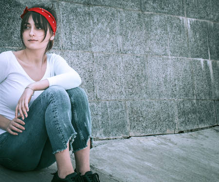 Beautiful girl in jeans and a white T-shirt with a red armband on the head, against the wall background, the concept of urban clothing and youth styleの写真素材