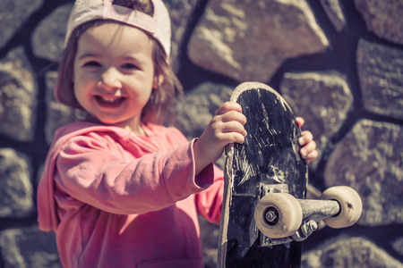 A fashionable little girl is holding a skateboard and playing outside, the beautiful emotions of a child, the concept of a carefree and fun-filled childhood. Children's skateboarding.の写真素材
