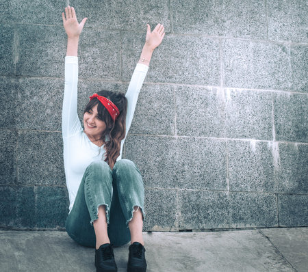 Beautiful girl in jeans and a white T-shirt with a red armband on the head, against the wall background, the concept of urban clothing and youth styleの写真素材