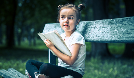 little cute girl is sitting on a bench and is reading a book in the park on a background of green natureの写真素材