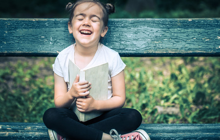 little cute girl is sitting on a bench and is reading a book in the park on a background of green natureの写真素材