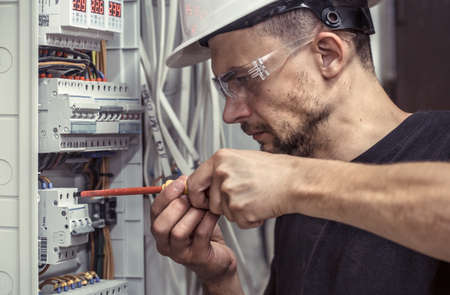 a male electrician works in a switchboard with an electrical connecting cable, connects the equipment with tools, the concept of complex work, space for textの写真素材