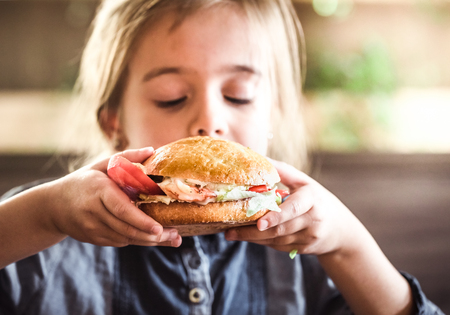 Little cute girl eating a sandwich in a cafe, concept of a children's fast food mealの写真素材