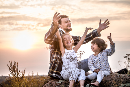 Happy loving family, dad playing with two little cute daughters at sunset sitting on a high beautiful mountain, the concept of family relationships and friendshipの写真素材