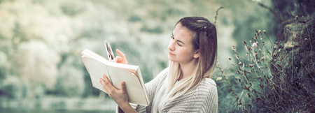 A young girl in a sweater reads a book against a background of nature, a concept of leisure and a hobbyの写真素材