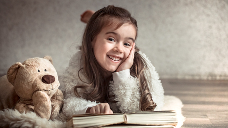 little girl reading a book with a Teddy bear on the floor in a cozy living room at home, beautiful emotions, the concept of relaxation and friendshipの写真素材