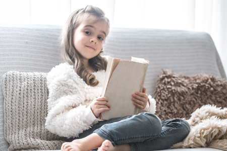 little girl reading a book on a comfortable sofa in a cozy living room at home, beautiful emotions, the concept of relaxationの写真素材