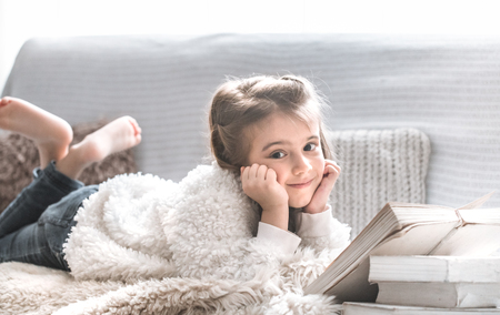little girl reading a book on a comfortable sofa in a cozy living room at home, beautiful emotions, the concept of relaxationの写真素材