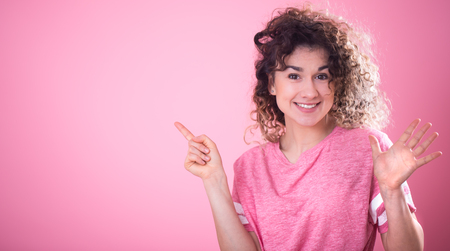 Portrait of a young cheerful girl with curly hair in a loose pink t-shirt shows finger to the side, isolated on a pink background, place for text, emotion conceptの写真素材