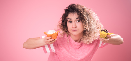 Portrait of a young curly-haired girl with cupcakes in her hands, in a loose pink t-shirt isolated on a pink background, a place for text, the concept of emotions and healthy eatingの写真素材