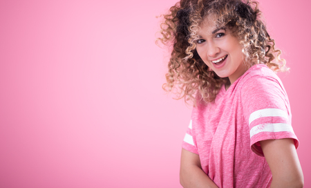 Portrait of young beautiful cheerful girl with curly hair and loose pink t-shirt isolated on pink background, place for text, woman concept and emotionの写真素材