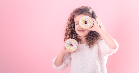 Portrait of a little smiling girl with curly hair and two appetizing donuts in her hands, closes her eyes with donuts, on a pink background, a place for textの写真素材