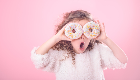 Portrait of a little surprised girl with curly hair, and two mouth-watering donuts in her hands, closes her eyes with donuts, on a pink backgroundの写真素材