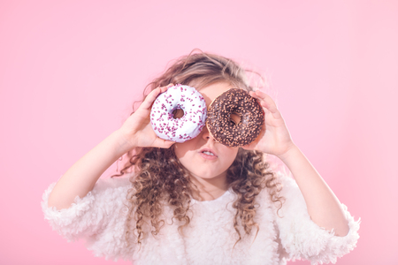 Portrait of a little surprised girl with curly hair, and two mouth-watering donuts in her hands, closes her eyes with donuts, on a pink backgroundの写真素材