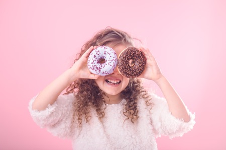 Portrait of a little smiling girl with curly hair and two appetizing donuts in her hands, closes her eyes with donuts, on a pink background, a place for textの写真素材