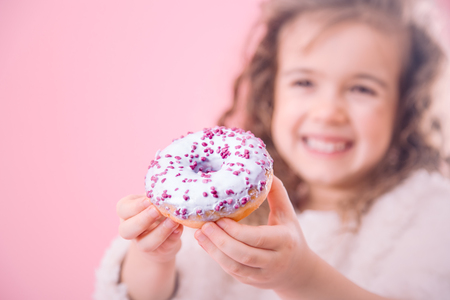 Portrait of a little smiling girl with curly hair and appetizing donuts in her hands, on a pink background, a place for textの写真素材