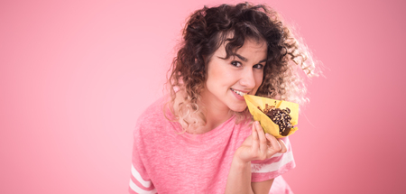 Portrait of a cheerful young curly-haired girl with cupcakes in her hands, in a loose pink t-shirt isolated on a pink background, a place for text, the concept of emotions and healthy eatingの写真素材