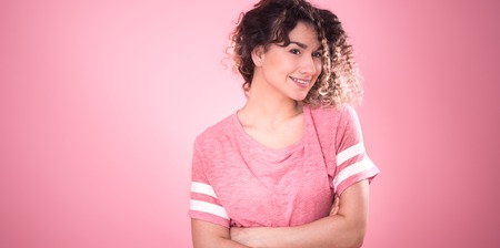 Portrait of young beautiful cheerful girl with curly hair and loose pink t-shirt isolated on pink background, place for text, woman concept and emotionの写真素材