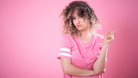Portrait of young surprised girl with curly hair in loose pink t-shirt shows finger to the side isolated on pink background, place for text, emotion conceptの写真素材
