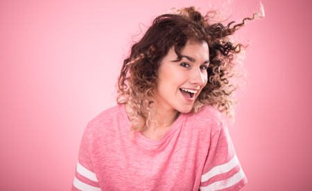 Portrait of young beautiful cheerful girl with curly hair and loose pink t-shirt isolated on pink background, place for text, woman concept and emotionの写真素材