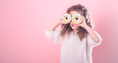 Portrait of a little surprised girl with curly hair, and two mouth-watering donuts in her hands, closes her eyes with donuts, on a pink backgroundの写真素材