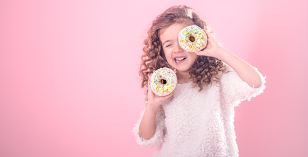 Portrait of a little smiling girl with curly hair and two appetizing donuts in her hands, closes her eyes with donuts, on a pink background, a place for textの写真素材