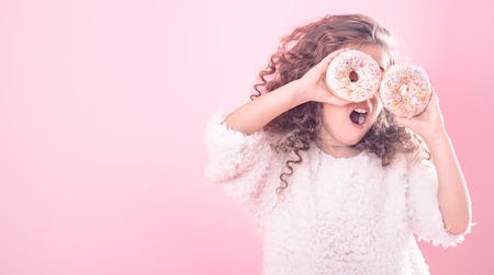 Portrait of a little curly girl with an open mouth and donuts in her hands, closes her eyes with donuts, on a pink background, a place for textの写真素材