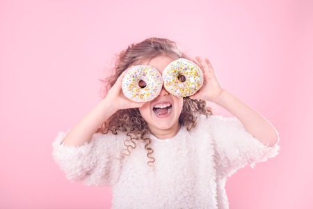 Portrait of a little smiling girl with curly hair and two appetizing donuts in her hands, closes her eyes with donuts, on a pink background, a place for textの写真素材