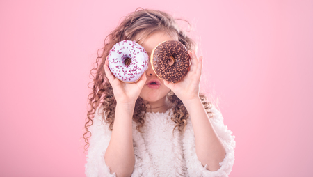 Portrait of a little surprised girl with curly hair, and two mouth-watering donuts in her hands, closes her eyes with donuts, on a pink backgroundの写真素材