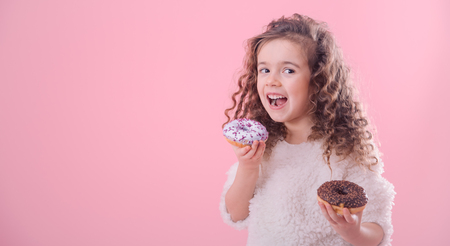 Portrait of a little joyful cute curly girl who eats donuts, on a pink background, a place for textの写真素材